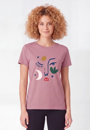 Vrouw met krullend haar die een mauve t-shirt draagt met abstracte gezichtsart met een maan, zon, lippen en groene bladvormen.