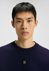 Ung man med kort svart hår iklädd en marinblå sweatshirt med en centrerad svart kvadrat HUGO-logotyp.