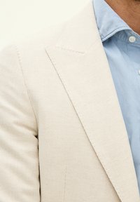 Ljusbeige strukturerad kavaj med slag, som matchas med en ljusblå skjorta med krage. Närbild visar sömnadsdetaljer och tygväv.