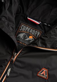 Veste noire Superdry avec fermeture éclair orange et blanche, écusson logo montagne et étiquette intérieure présentant un design de montagne au coucher du soleil ainsi qu'une taille moyenne.