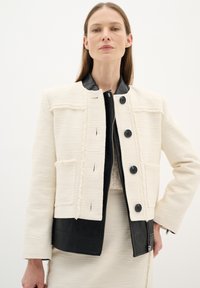 Giacca bianca testurizzata con accenti in pelle nera, caratterizzata da un taglio dritto, colletto rotondo e tasche frontali, chiusa con grandi bottoni.