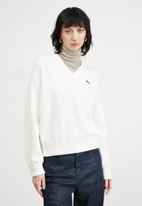 Witte v-hals sweater van een zachte stof, met een groen logo op de borst, gecombineerd met een beige coltrui eronder. Donkerblauwe spijkerbroek.