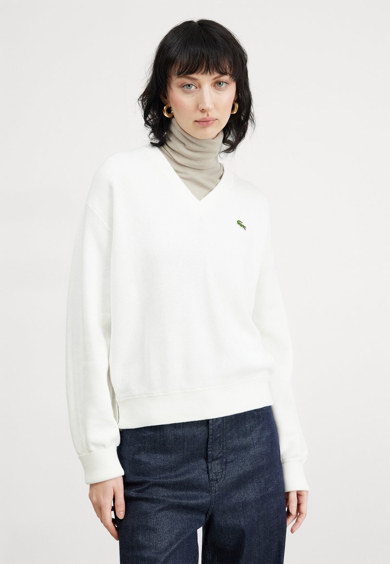 Witte v-hals sweater van een zachte stof, met een groen logo op de borst, gecombineerd met een beige coltrui eronder. Donkerblauwe spijkerbroek.