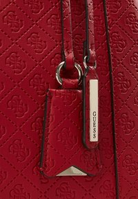 Rode synthetische leren handtas met een reliëfpatroon, zilverkleurige hardware en een label met de merknaam "Guess." Gestructureerde vorm en een textuurfinish.