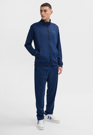 Navy blue tracksuit made of smooth fabric, featuring a zippered jacket with stand-up collar and matching pants, worn with white and blue sneakers.