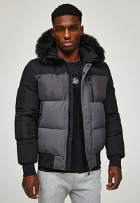 Black and gray padded jacket with a fur-lined hood, featuring a zip closure, side pockets, and a logo on the chest.