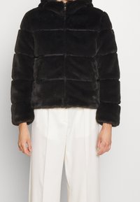 Svart konstpälsjacka med huva, vadderad design och horisontella sömmar. Har en dragkedja fram och vida ärmar, stylar med vita byxor.