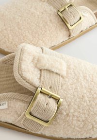 Pantoufles beiges avec un dessus en polaire doux et texturé et une lanière en tissu côtelé. Comprend une boucle dorée et une semelle plate en caoutchouc beige.