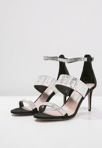 Sandales à talons hauts noires avec des brides en simili-cuir crocodile argenté brillant et une seule bride de cheville. Matériau en daim doux, design à bout ouvert.