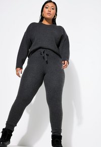 Grauer Strickpullover mit herunterhängenden Schultern und einem Kordelzugbund, kombiniert mit passenden, enganliegenden Jogginghosen mit elastischen Bündchen.