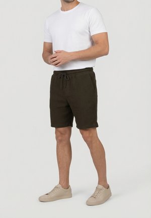 Homme portant un t-shirt blanc à manches courtes, un short à cordon de serrage olive foncé et des baskets beiges, debout devant un fond uni.