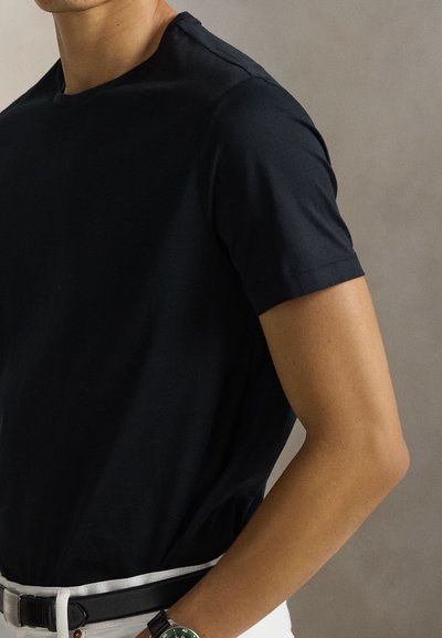 Άνδρας με μαύρο κοντομάνικο πουκάμισο, λευκό παντελόνι με μαύρη ζώνη και μαύρο ρολόι με πράσινο καντράν, στέκεται μπροστά σε απλό φόντο.