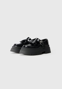 Chaussures à plateforme noires avec une finition brillante, dotées d'une semelle en caoutchouc épaisse, de sangles croisées et d'une boucle sur le côté pour la fermeture. Design de bout plat.