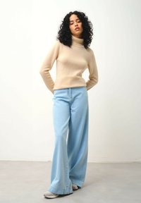 Beige Rollkragenpullover und hellblaue weit geschnittene Jeans. Der Pullover ist figurbetont; die Jeans haben einen leicht ausgestellten Saum und subtile Nähte.