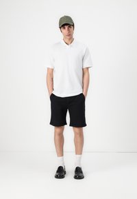 White polo shirt, black shorts, green cap, black loafers, white socks. Model stands hands in pockets against plain background.