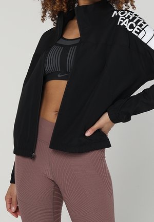 Kobieta w czarnej kurtce North Face na czarnym sportowym biustonoszu Nike i lawendowych teksturowanych legginsach, z jedną ręką na biodrze.