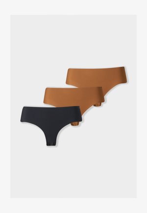Femme aux longs cheveux foncés portant un soutien-gorge noir et des sous-vêtements marron, debout pieds nus devant un fond clair et uni.