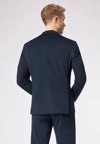 Uomo di spalle che indossa un giubbotto di giacca a coste blu navy e pantaloni abbinati su uno sfondo chiaro e semplice.