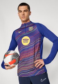Nike majica s dugim rukavima ima plavu osnovu s višebojnim valovitim linijama, žuti Spotify logo i grb FC Barcelone. Igrač drži crno-bijelu nogometnu loptu s crvenim detaljima.