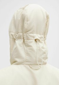 Polstret hættetrøje i lys beige. Har en snorafslutning ved halsen, med tekstureret ydre stof og syninger.
