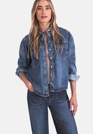 Femme portant une veste en denim bleue sur un haut rose clair, associée à un jean bleu, posant avec une main dans la poche devant un fond blanc.