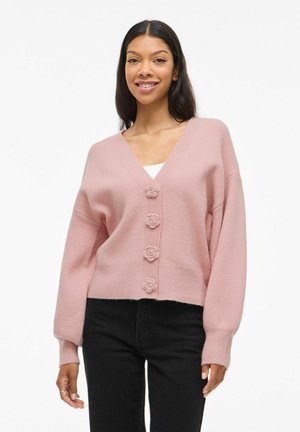 Cárdigan rosa claro con escote en V, cuatro botones en forma de rosa, puños acanalados y un diseño corto, combinado con pantalones negros. Textura suave.