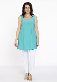 Ein ärmelloses türkises Oberteil mit A-Linien-Form, kombiniert mit weißen Hosen. Das Outfit wird durch eine runde goldene Halskette mit Anhänger ergänzt.