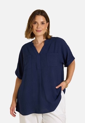 Woman with shoulder-length brown hair wearing gold hoop earrings, a loose navy blue shirt with pockets, white pants, and gold bracelets.