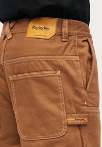 Καφέ παντελόνι με μεγάλη πλαϊνή τσέπη, λεπτομερείς ραφές και ετικέτες Replay Inc. στη μέση και στον ιμάντα της τσέπης.