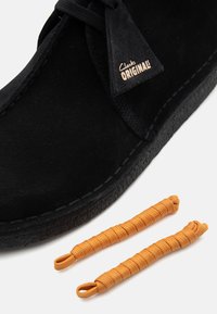 Black suede boot with a textured surface and black rubber sole. Includes two orange laces with a spiral texture. Brand label visible.