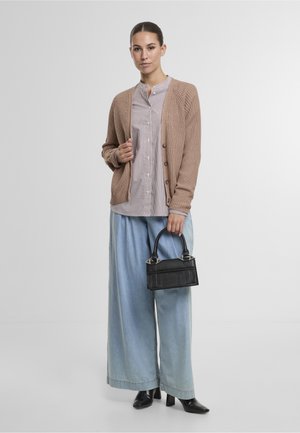 Sieviete, kura valkā bēšas krāsas kardiganu pārsvītrotas blūzes virspusē, gaiši zilas plaša piegriezuma džinsas un melnas augstpapēžu zābaki, turot nelielu melnu rokassomiņu pelēka fona priekšā.