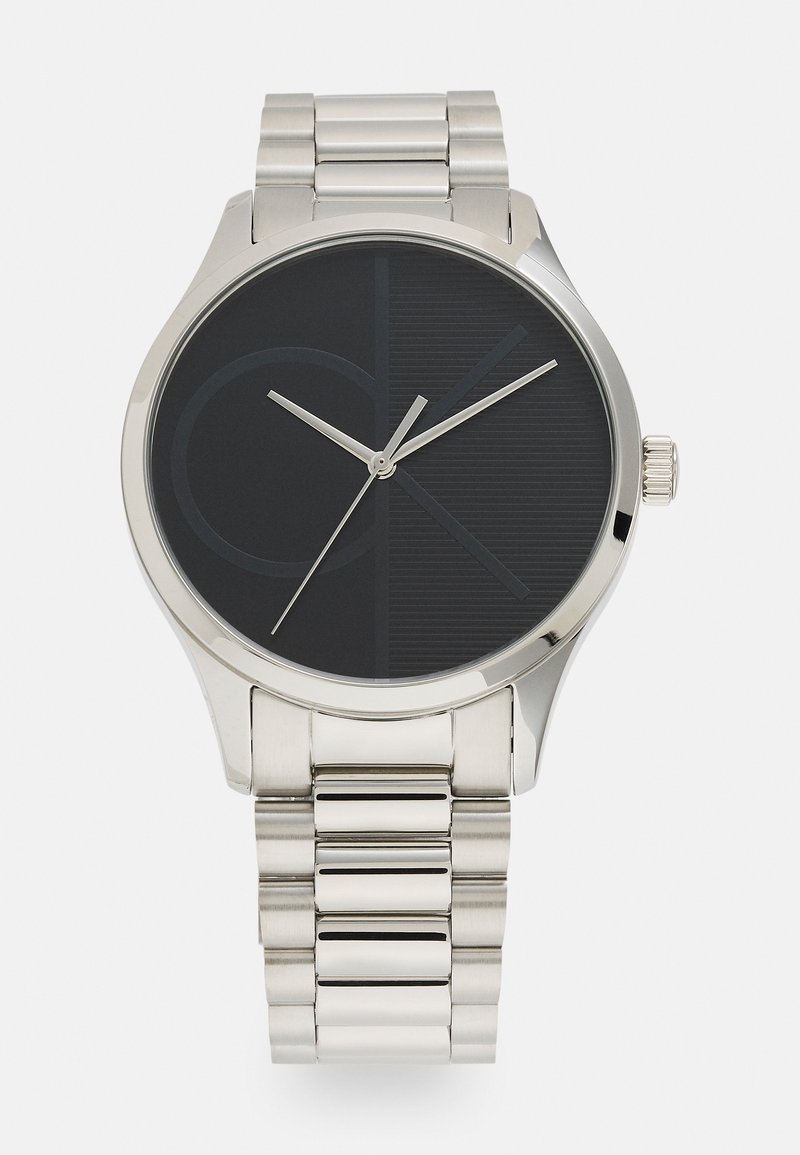 Zilveren roestvrijstalen horloge met een zwarte wijzerplaat. Kenmerkt zich door een minimalistisch ontwerp met CK-logo, dunne wijzers en een gestructureerde achtergrond.
