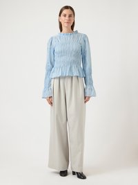 Γυναίκα που στέκεται, φοράει ανοιχτό μπλε μπλούζα με μακριά μανίκια και ρυτιδώσεις, μπεζ παντελόνι με φαρδιά πόδια και μαύρα παπούτσια με τακούνι, μπροστά από ένα απλό φόντο.