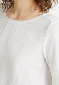 Weißes Langarmoberteil mit rundem Ausschnitt, glattem Material, subtilem Glanz und kontrastierenden grauen Akzenten entlang der Kragennaht.