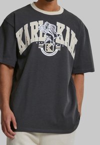 Gray oversized t-shirt featuring a white and beige graphic print, with text and an eagle design, along with a contrasting ribbed collar.