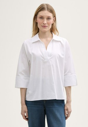 Blouse en coton blanc avec un col en V et des manches trois-quarts, présentant une coupe décontractée et une texture lisse. Associée à un jean en denim bleu.