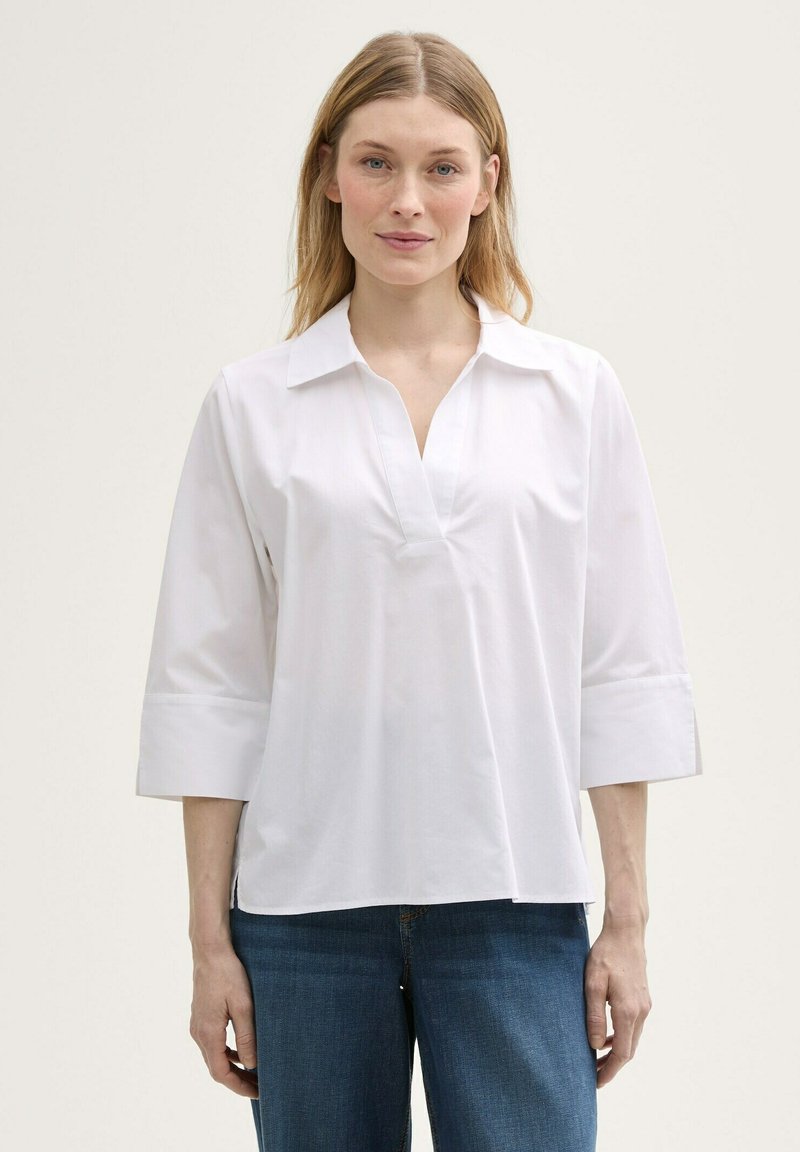 Blouse en coton blanc avec un col en V et des manches trois-quarts, présentant une coupe décontractée et une texture lisse. Associée à un jean en denim bleu.