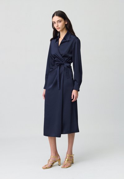 Vestido envolvente de satén azul marino con mangas largas, cuello y cinturón autoadherente. Combinado con sandalias doradas de tacón con correas y hebilla en el tobillo.