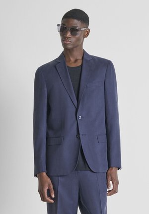 Man in een marineblauw pak, een zwarte trui en donkere zonnebril, staande tegen een effen lichtgrijze achtergrond.