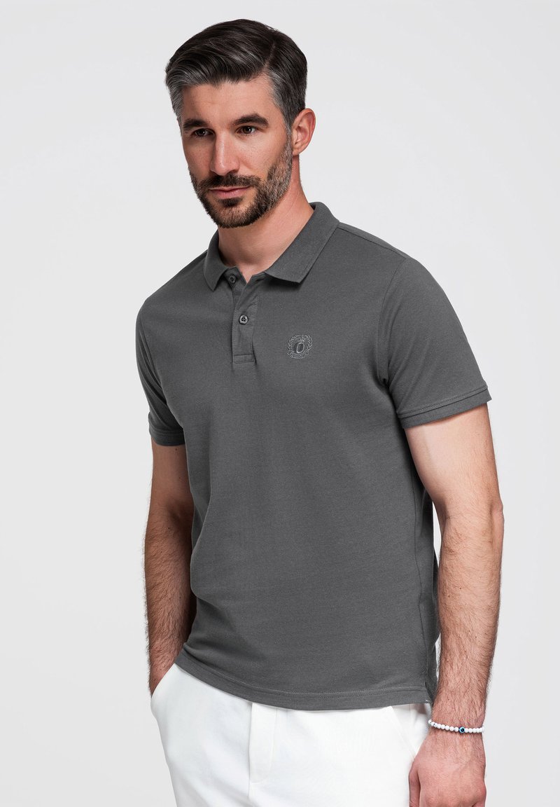 Man met kort donker haar en baard draagt een grijs poloshirt en witte broek, staat met één hand in zijn zak tegen een effen achtergrond.