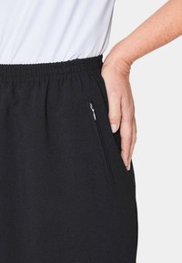 Schwarze Jogginghose mit elastischem Bund, seitlichem Reißverschlussfach und glatter Textur. Hand auf der Tasche platziert.