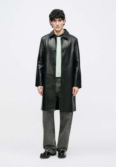 Abrigo negro de cuero con cierre de botones en la parte delantera, mangas largas y silueta recta, usado sobre una camiseta verde claro y jeans grises.