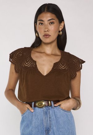 Donna con capelli lunghi e scuri che indossa orecchini a cerchio dorati, un top marrone con spalle di pizzo, jeans blu, una cintura con una grande fibbia ovale e braccialetti.