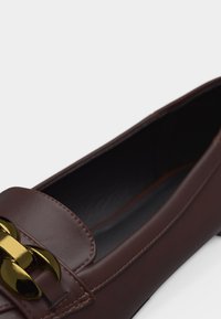 Mocassin en cuir marron foncé avec une large chaîne en métal doré sur le dessus et des coutures lisses le long des bords.