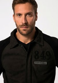 Sort jakke med tekstureret overflade, der har en krave og en frontlynlås, udsmykket med numrene "849" og "FIGHTER PILOT" i broderede lapper.
