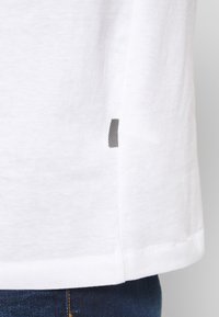 Hvid bomulds-t-shirt med en glat tekstur, der har et sort stribet mærke på sidesømmen. Kantene er let buede.