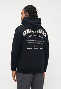 Man with dreadlocks wearing a black hoodie with "ORIGINALS INTERNATIONAL Connecting Creativity Across Distances" text on the back.