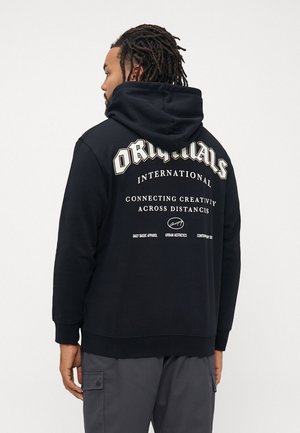 Mann med dreadlocks iført en svart hettegenser med teksten "ORIGINALS INTERNATIONAL Connecting Creativity Across Distances" på baksiden.