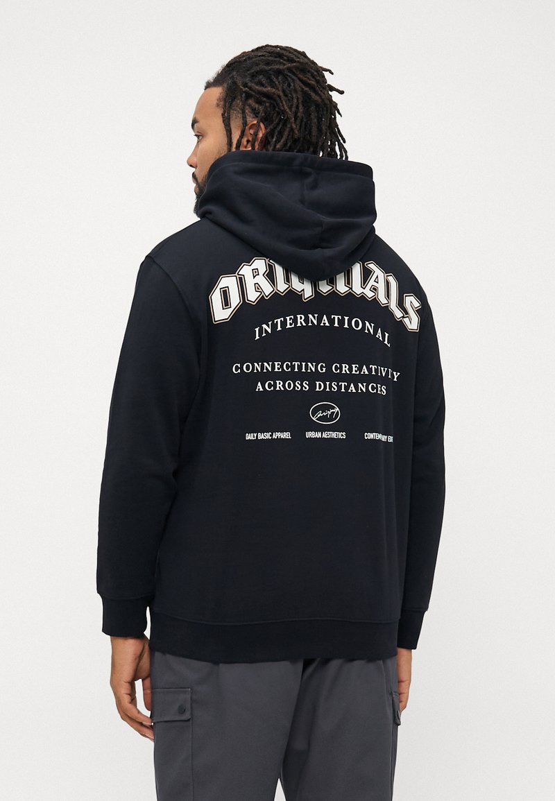 Man with dreadlocks wearing a black hoodie with "ORIGINALS INTERNATIONAL Connecting Creativity Across Distances" text on the back.