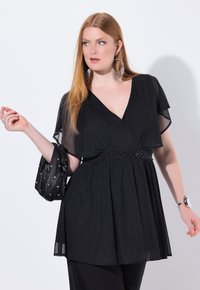 Top nero avvolgente con maniche trasparenti, dettaglio in perline in vita, tessuto fluido e leggero splendore. Modella che indossa orecchini statement e un bracciale.
